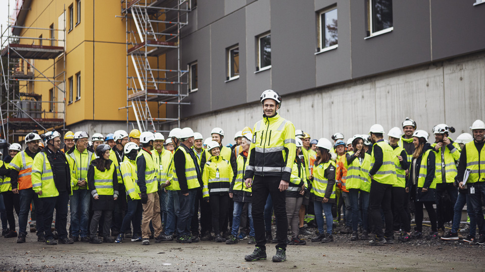 Projektledare från Specialfastigheter med många byggnadsarbetare bakom sig framför ett nybyggt typhus